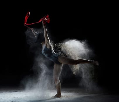 beautiful caucasian woman in a black bodysuit with a sports figure dancing with red ribbon on a black background, gymnastic exercises in flying white flour