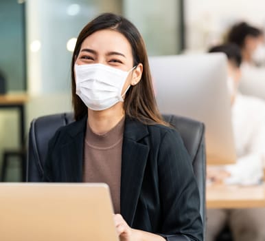 Portrait of asian office employee businesswoman wear protective face mask work in new normal office with interracial colleague in background as social distance practice prevent coronavirus COVID-19.