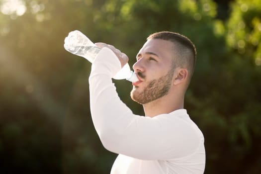 The sporty young guy is drinking water from bottle for hydrating outdoors in nature after recreation.