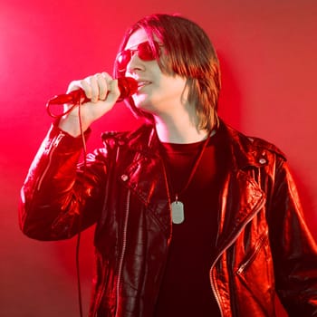 The young guy in black leather jacket is singing rock music on red illuminated stage.