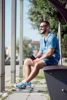 The smiling handsome young guy with earphones is sitting and listening music outdoors.