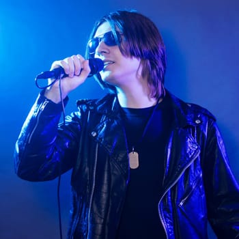 The young guy in black leather jacket is singing rock music on blue illuminated stage.