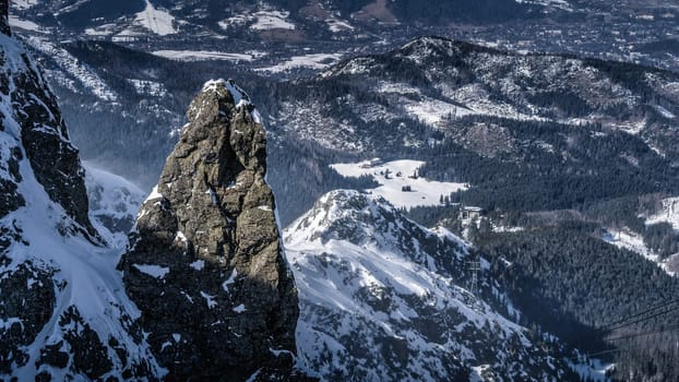 Large pointy rock covered in snow, highlighted by sunlight with winter shelter and Zakopane village below in the valley, Bukowina Tatrzanska, Poland