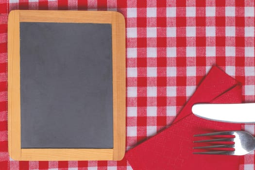 Food recipe template. Empty wooden black board and knife and fork on a red white checkered tablecloth. For your food and product display montage. Copy space.