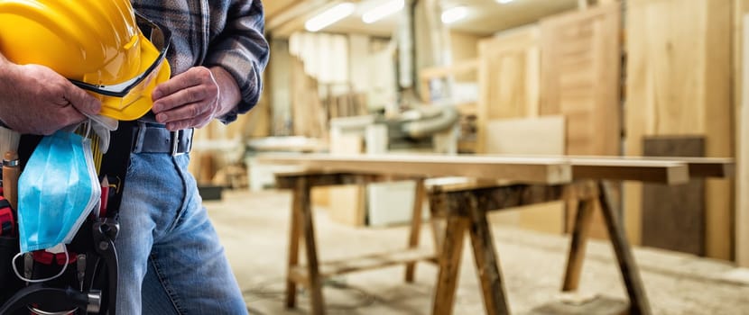 Carpenter in the carpentry workshop, holds helmet, goggles, leather gloves and surgical mask in hand to prevent Coronavirus infection. Preventing Pandemic Covid-19 at the workplace.