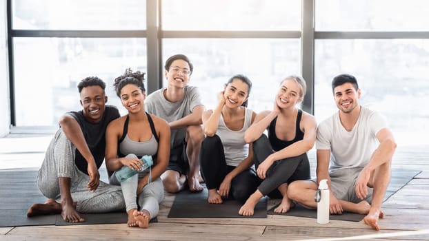 Happy sporty people posing in fitness studio after yoga training, sitting on mats and smiling, enjoying healthy lifestyle