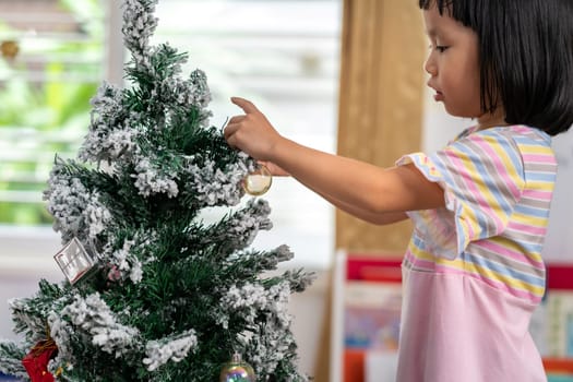 Little asian girl decorating a Christmas tree in her living room with christmas ornament preparing for season greeting of Merry Christmas and Happy Holidays.