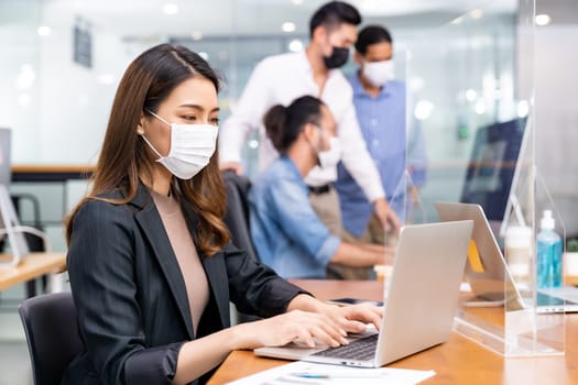 Portrait of asian office employee businesswoman wear protective face mask work in new normal office with interracial team in background as social distance practice prevent coronavirus COVID-19.