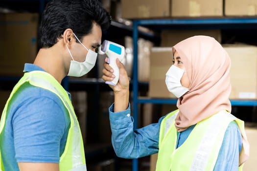 Islam asian warehouse worker taking temperature to worker before getting in factory after reopening from city lockdown from COVID-19 coronavirus pandemic in distribution center environment. Side view.