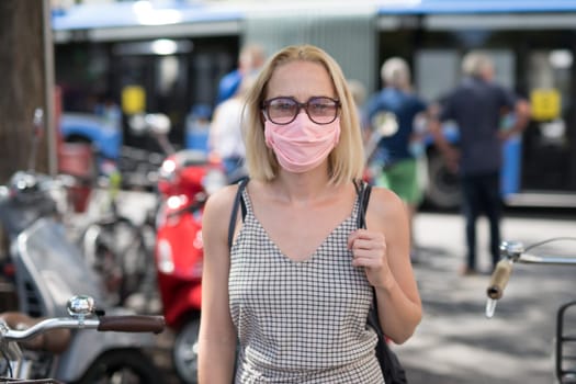 Portrait of casual yound woman walking on the street wearing protective mask as protection against covid-19 virus. Incidental people on the background.