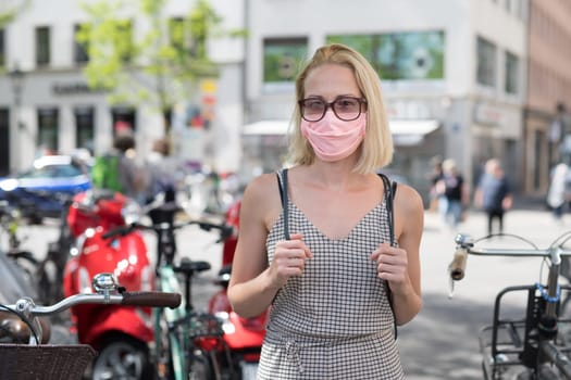 Portrait of casual yound woman walking on the street wearing protective mask as protection against covid-19 virus. Incidental people on the background.