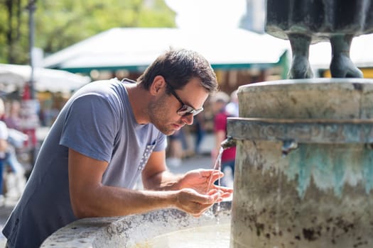 Thirsty young casual cucasian man drinking water from public city fountain on a hot summer day.