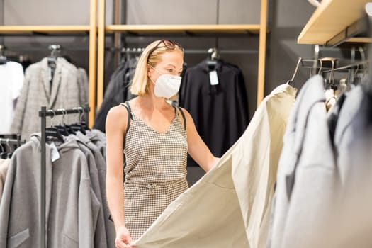 Fashionable woman wearing protective face mask shopping clothes in reopen retail shopping store. New normal lifestyle during corona virus pandemic.