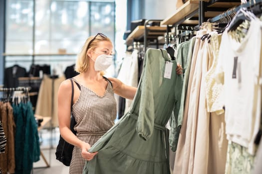 Fashionable woman wearing protective face mask shopping clothes in reopen retail shopping store. New normal lifestyle during corona virus pandemic.