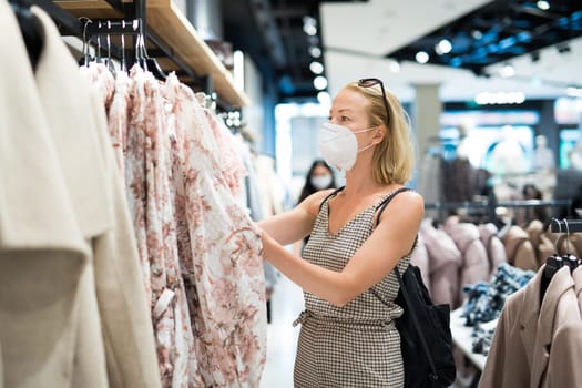 Fashionable woman wearing protective face mask shopping clothes in reopen retail shopping store. New normal lifestyle during corona virus pandemic.