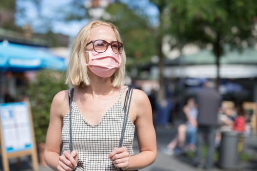 Portrait of casual yound woman walking on the street wearing protective mask as protection against covid-19 virus. Incidental people on the background.