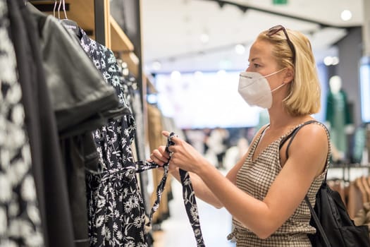 Fashionable woman wearing protective face mask shopping clothes in reopen retail shopping store. New normal lifestyle during corona virus pandemic.