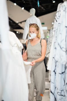 Fashionable woman wearing protective face mask shopping clothes in reopen retail shopping store. New normal lifestyle during corona virus pandemic.