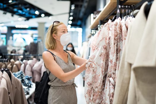 Fashionable woman wearing protective face mask shopping clothes in reopen retail shopping store. New normal lifestyle during corona virus pandemic.