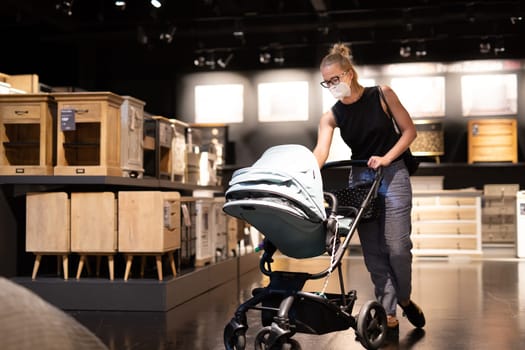 Young mom with newborn in stroller shopping at retail furniture and home accessories store wearing protective medical face mask to prevent spreading of corona virus. New normal during covid epidemic.