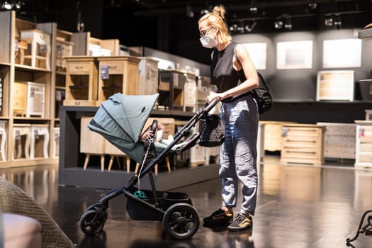 Young mom with newborn in stroller shopping at retail furniture and home accessories store wearing protective medical face mask to prevent spreading of corona virus. New normal during covid epidemic.