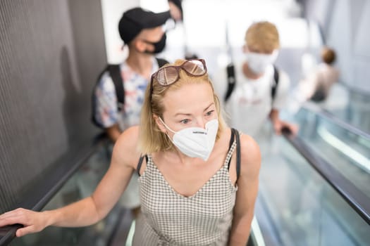 Portrait of casual yound woman uses escalators in the department store wearing protective mask as protection against covid-19 virus. Incidental people on the background.