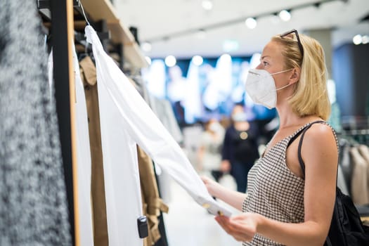 Fashionable woman wearing protective face mask shopping clothes in reopen retail shopping store. New normal lifestyle during corona virus pandemic.