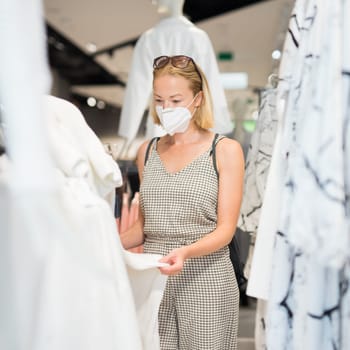 Fashionable woman wearing protective face mask shopping clothes in reopen retail shopping store. New normal lifestyle during corona virus pandemic.