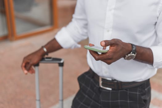 Black man with luggage bag standing on city street using smartphone, wearing formal style clothes.