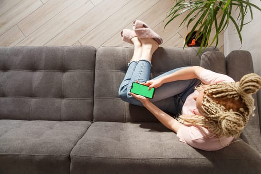 Woman with blond canecalons sitting on sofa and using smartphone with green screen for your image.