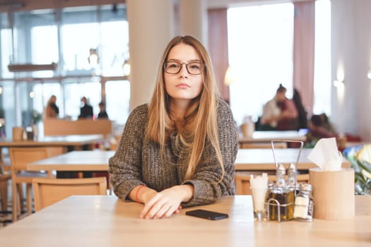 Portrait of cute freelancer girl sitting in a cafe. She wears brown hipster eyeglasses