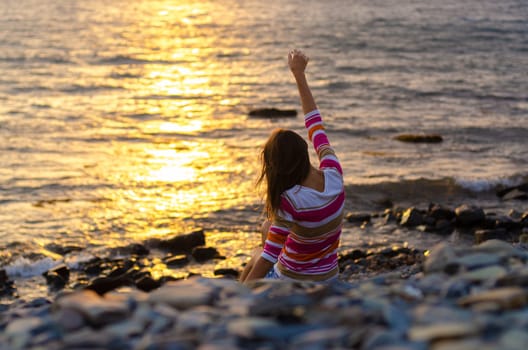 A girl sits on the seashore and throws stones into the water, raised her hands, evening, sunset