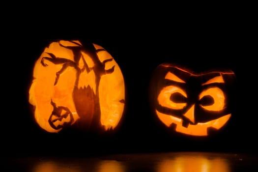 Two intricately carved halloween pumpkins lit by candle tea lights, one is of a scary face, and the other of terrifying trees and another pumpkin