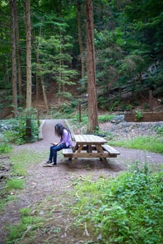 Young caucasian man with long hair and a purple shirt sits at a picnic table along an idyllic hiking trail to look at his phone.