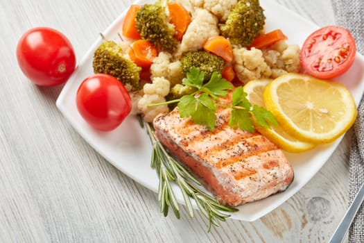 Grilled salmon fillet  garnished with vegetables, lemon, tomato, herbs lying on plate on wooden background. Top view. Healthy food. Keto diet.