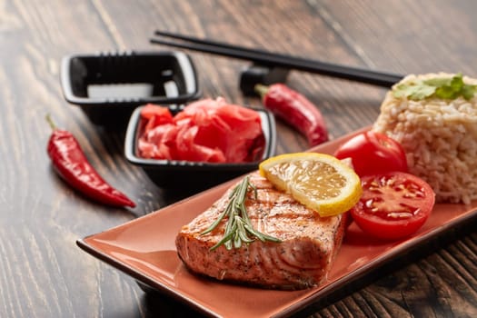 Grilled salmon fillet with rice, pickled ginger, soy sauce, tomato, lemon and herbs on dark wooden background.  Healthy food.