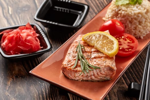 Grilled salmon fillet with rice, pickled ginger, soy sauce, tomato, lemon and herbs on dark wooden background.  Healthy food.