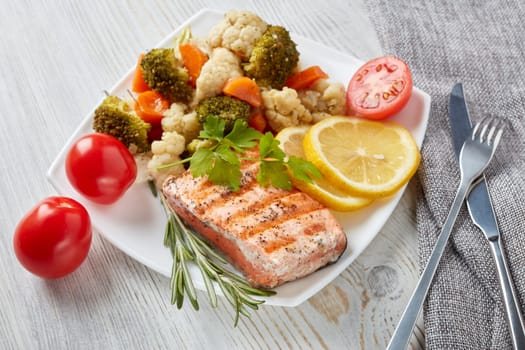 Grilled salmon fillet  garnished with vegetables, lemon, tomato, herbs lying on plate on wooden background. Top view.Healthy food. Keto diet.