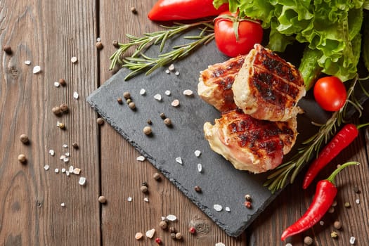 Bacon-wrapped grilled chicken (turkey)  fillet with fresh vegetables and spices on stone cutting board on wooden background. Top view with copy space