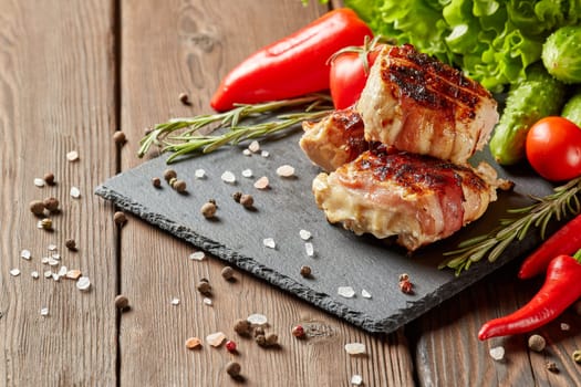 Bacon-wrapped grilled chicken (turkey) fillet with fresh vegetables and spices on stone cutting board on wooden background