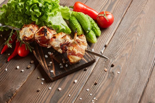 Shashlik from chicken with bacon. Grilled chicken (turkey) fillet on skewer and fresh vegetables with spices on stone cutting board on wooden background