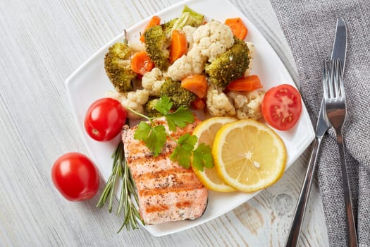 Grilled salmon fillet  garnished with vegetables, lemon, tomato, herbs lying on plate on wooden background. Top view.Healthy food. Keto diet.