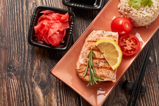 Grilled salmon fillet with rice, pickled ginger, soy sauce, tomato, lemon and herbs on dark wooden background. Top view. Healthy food.