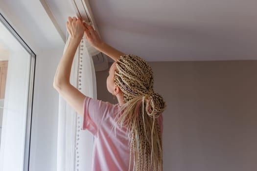 Young blonde woman hanging up curtains at the window