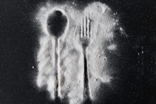 White sugar on black background with silhouette of spoon and fork.