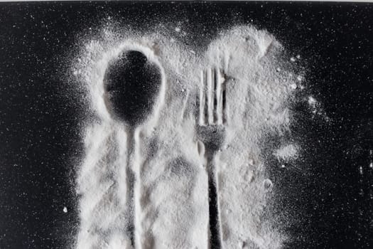 White sugar on black background with silhouette of spoon and fork.