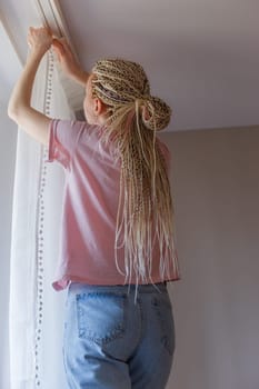Young blonde woman hanging up curtains at the window