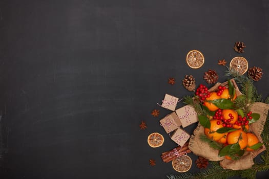 Festive still life: berries, mandarins with leaves on wooden tray with rough napkin from burlap, gift boxes, spices and fir branches on dark background. Top view with copy space.  Christmas. New Year