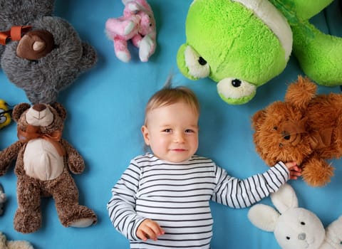 Happy one year old boy lying with many plush toys on blue blanket