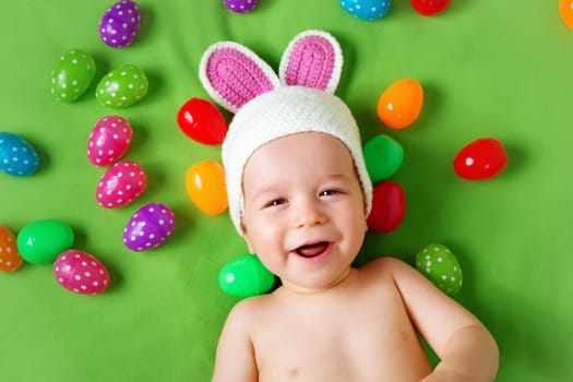 Baby boy in bunny hat lying on green blanket with multicolored easter eggs